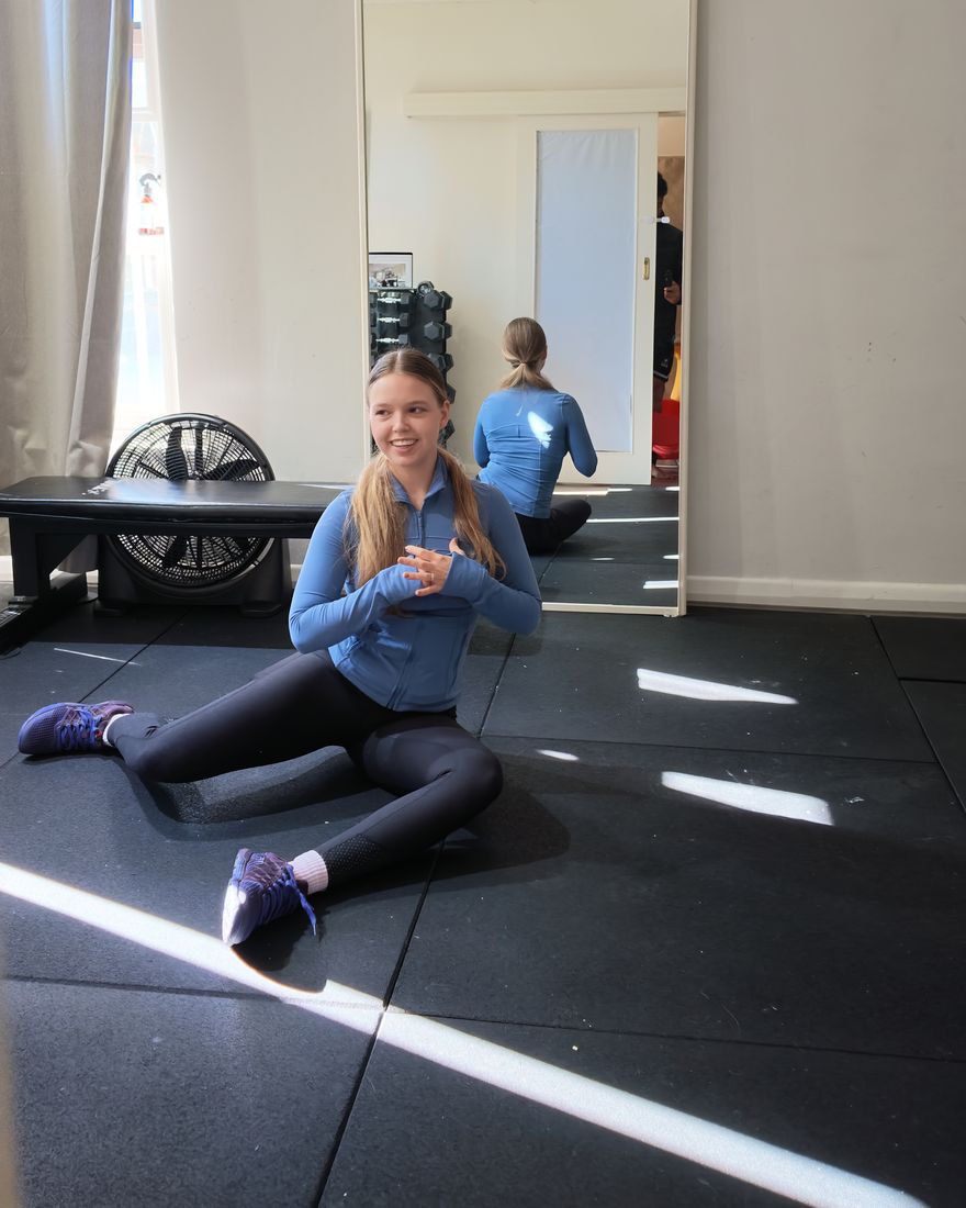 Liubov demonstrating a seated hip mobility exercise on a gym mat